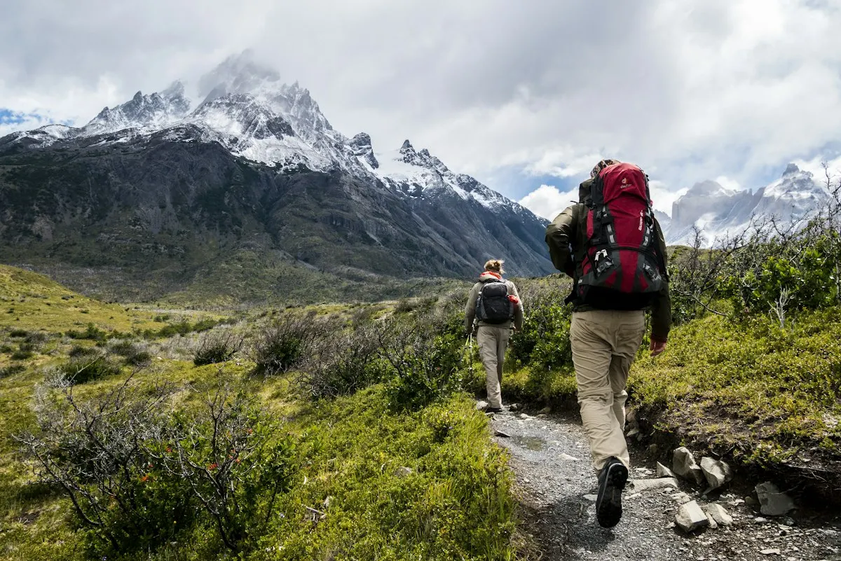 Two hikers with backpacks on a mountain trail below snow-capped peaks