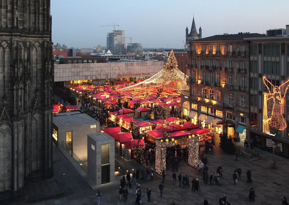 Red-roofed Christmas market stalls lit up in the square beside Cologne Cathedral at dusk
