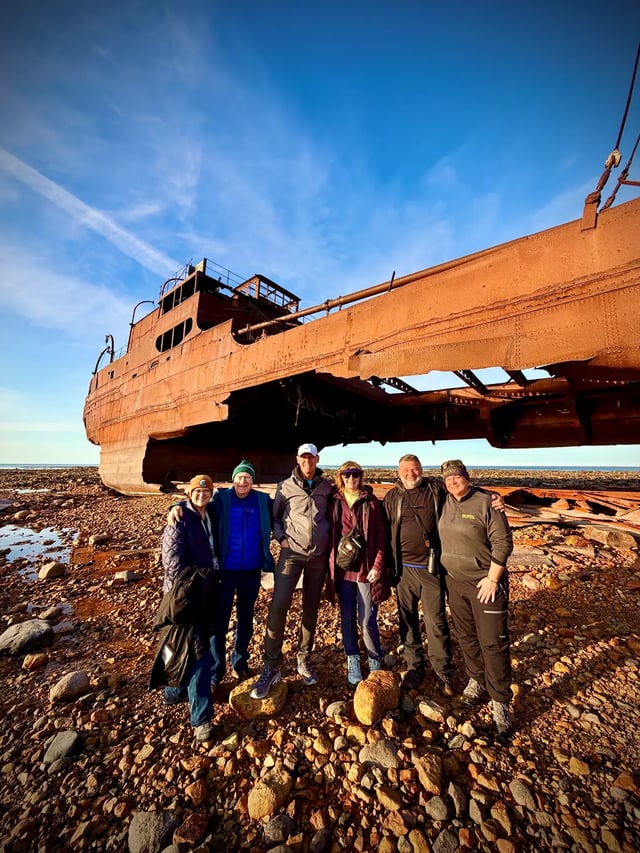 Journey with Erika travelers beneath the rusted Ithaca shipwreck in Churchill with Hudson Bay behind them