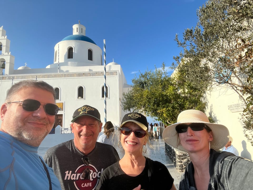 Four Journey with Erika travelers posing for a selfie on a paved walkway in Oia on Santorini, with the blue-domed Panagia Platsani church behind them