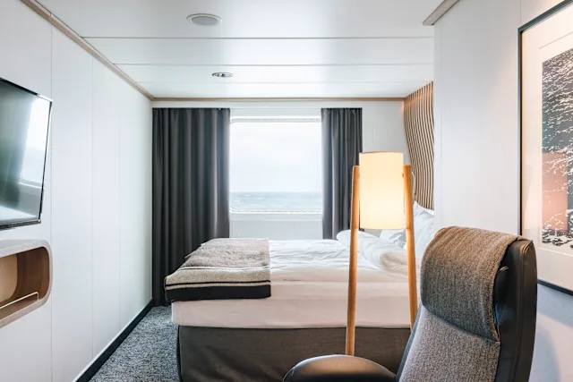 A bright ocean-view cabin with a bed, armchair and a window overlooking the sea