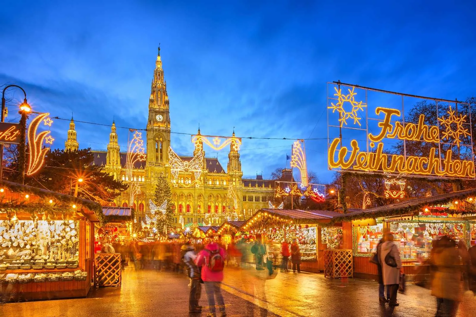 Vienna Christmas market glowing at dusk in front of the illuminated Rathaus, with a lit Christmas tree and a 'Frohe Weihnachten' light sign