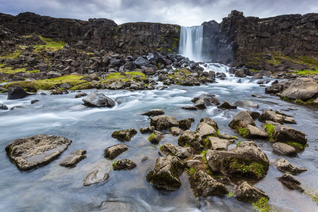 A waterfall pouring over a basalt cliff above a boulder-strewn river in Iceland