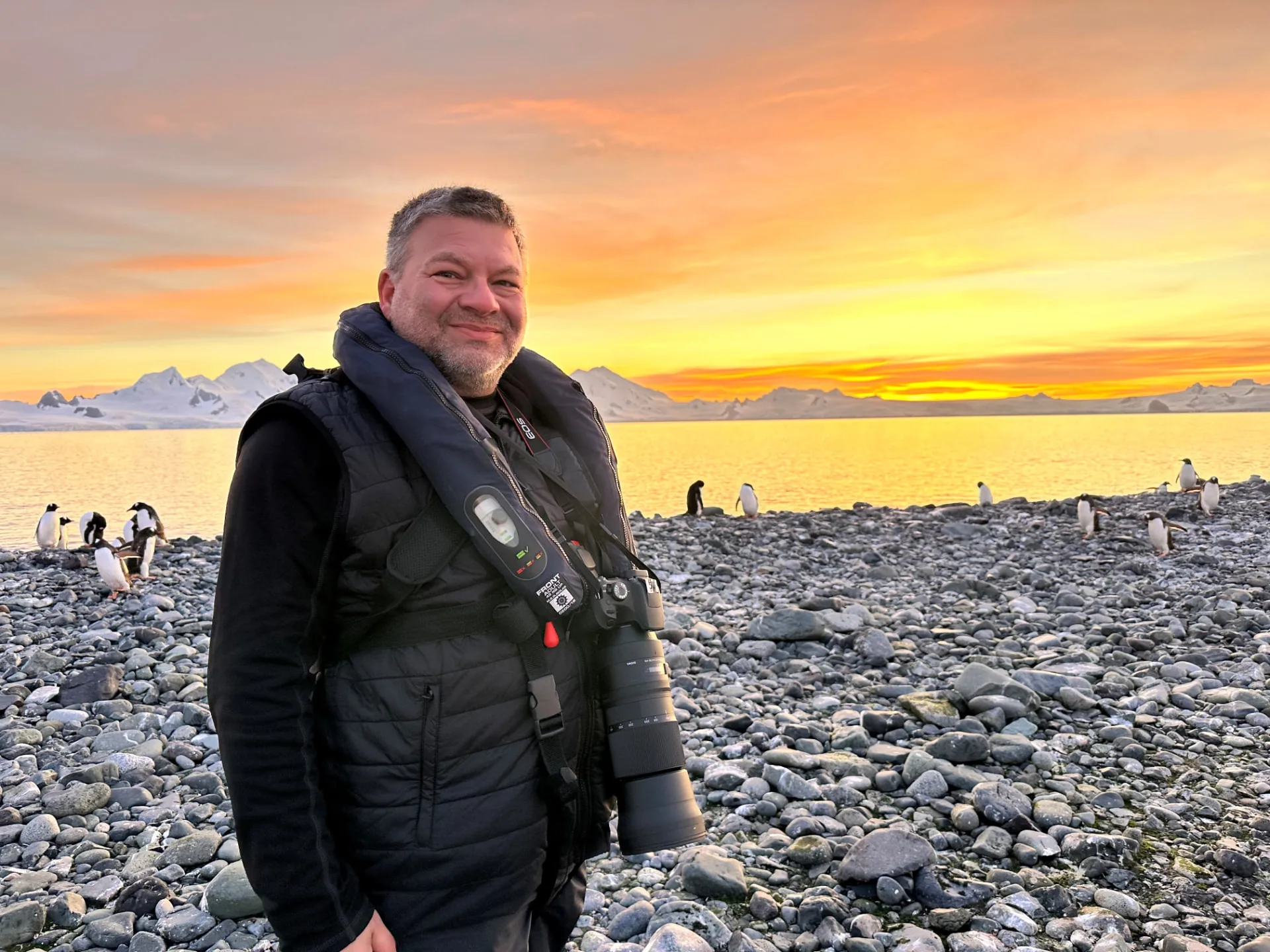Chris stands on a pebble beach among penguins at sunset in Antarctica, with a camera and snow-covered mountains behind him