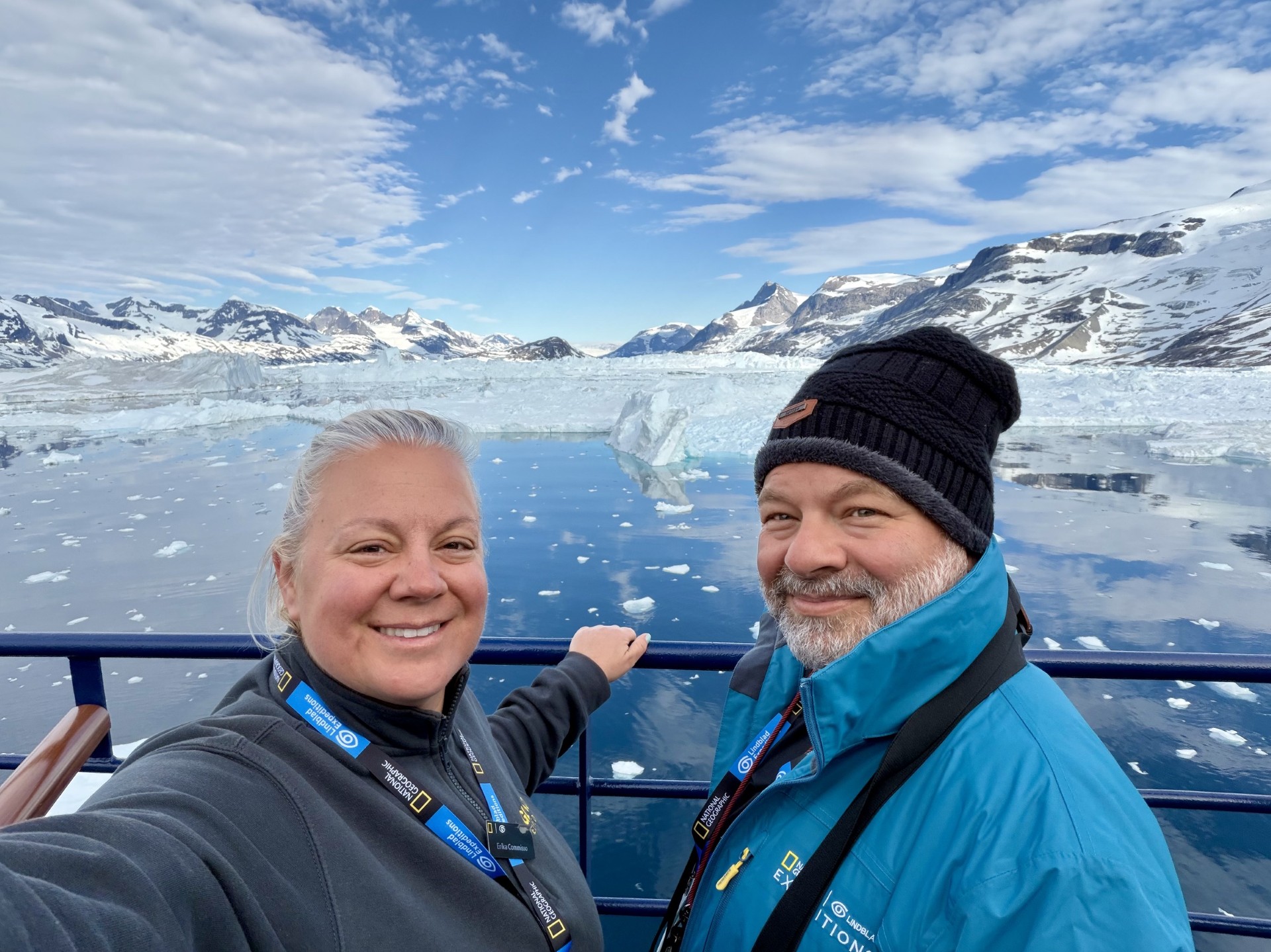 Erika and Chris Commisso smiling on the ship deck in front of icebergs and snow-capped mountains in Greenland