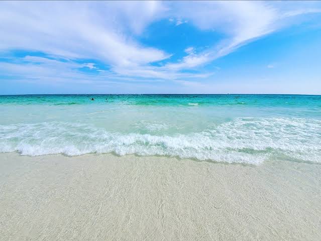 Turquoise waves rolling onto a white-sand beach under a bright blue sky with wispy clouds