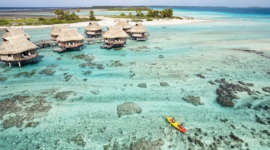 Aerial view of thatched overwater bungalows on a shallow turquoise lagoon, with two people kayaking in front