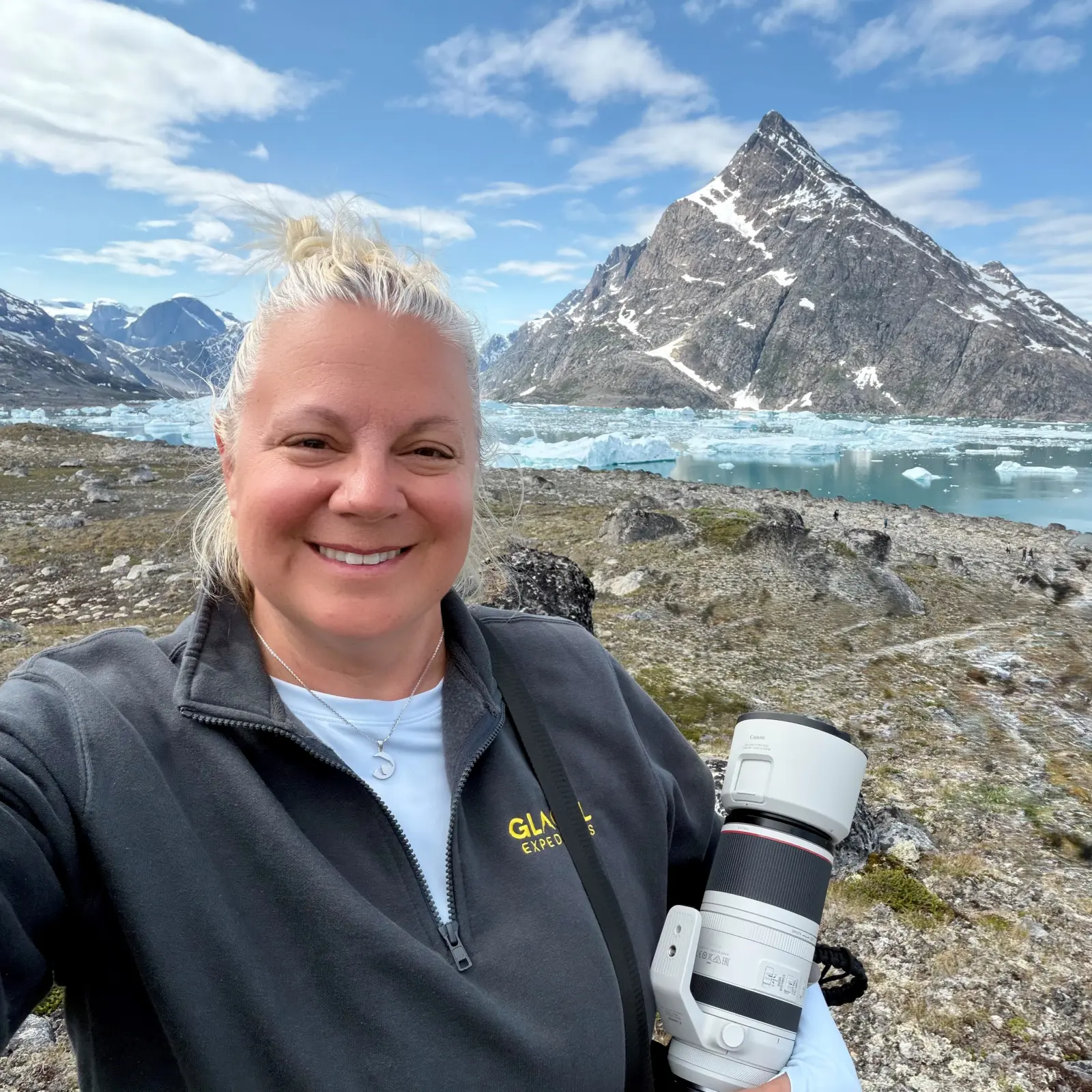 Erika Commisso, founder of Journey with Erika, smiling with a camera in front of icebergs and snow-dusted mountains in Greenland