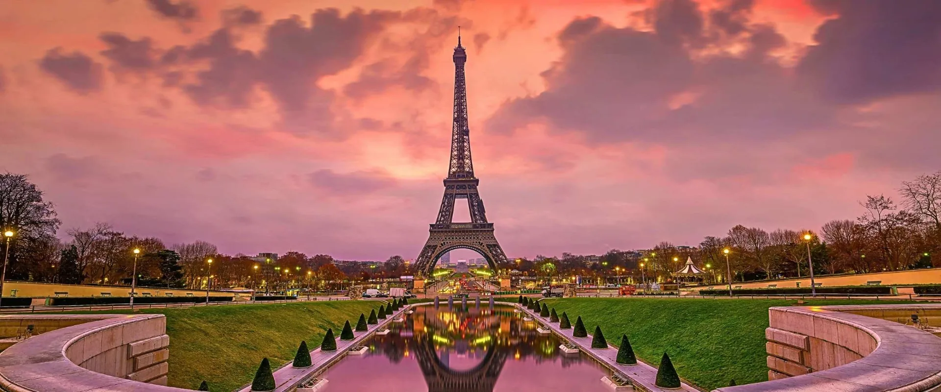 The Eiffel Tower seen from the Trocadéro at sunset, reflected in the long fountain pool below a pink and purple sky