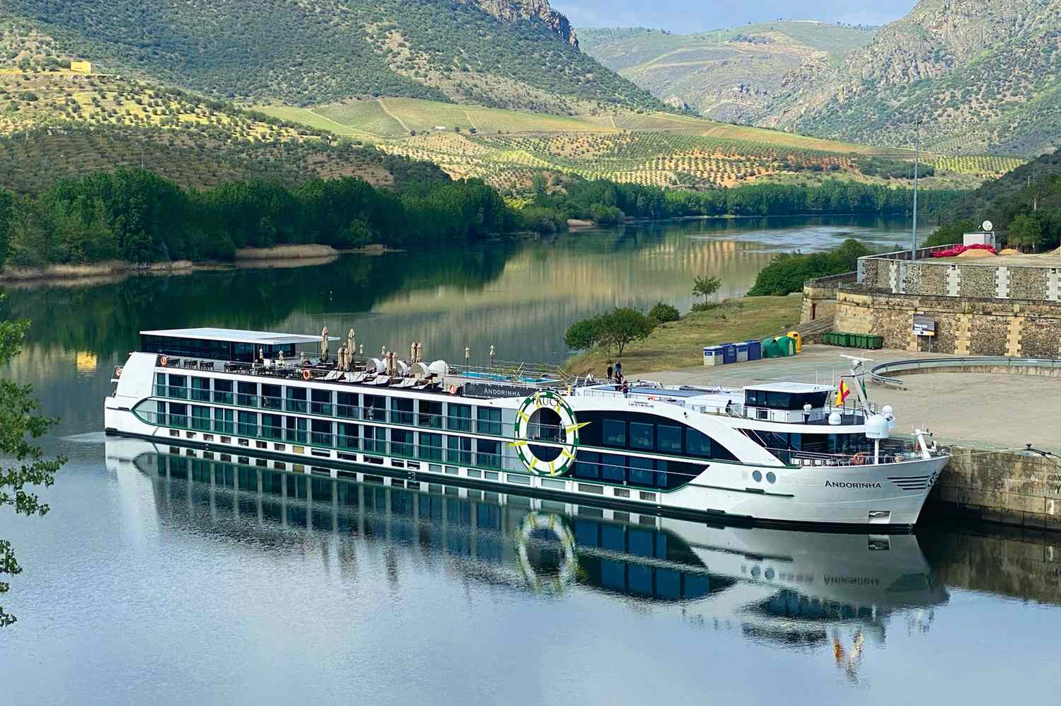 A river cruise ship docked on a calm river below terraced hillsides and a stone wall