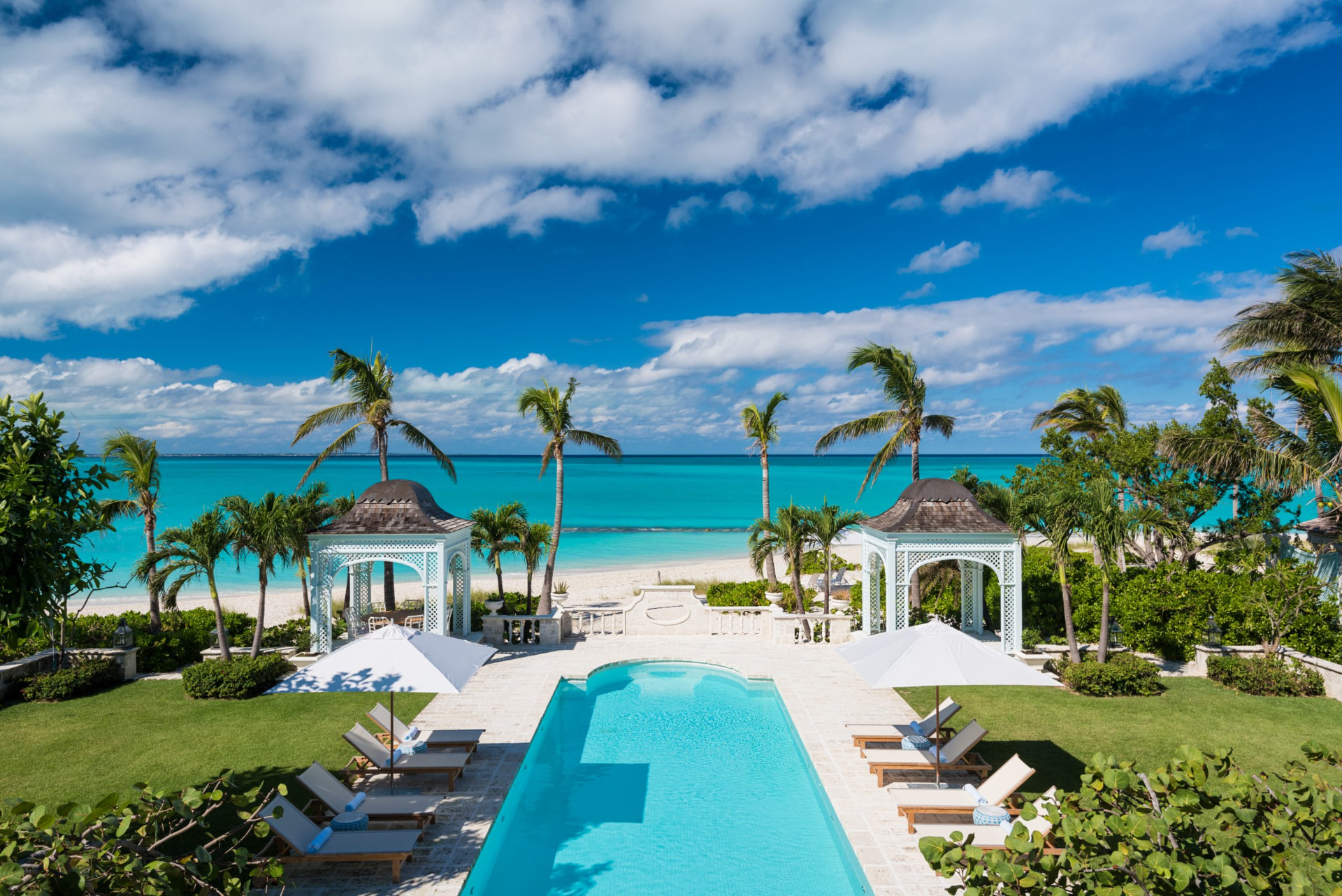 A turquoise resort pool lined with loungers and umbrellas between palm trees and white gazebos, opening onto a white-sand beach