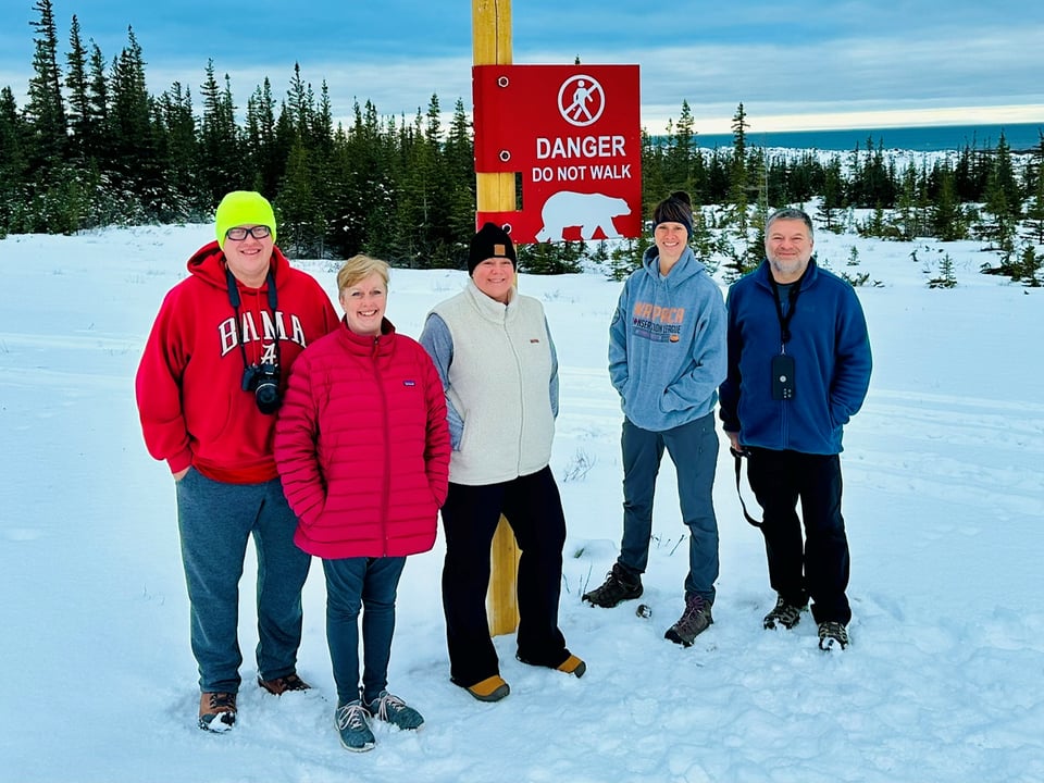 Journey with Erika travelers in winter coats beside a red polar bear danger sign in the snow, with spruce forest behind them