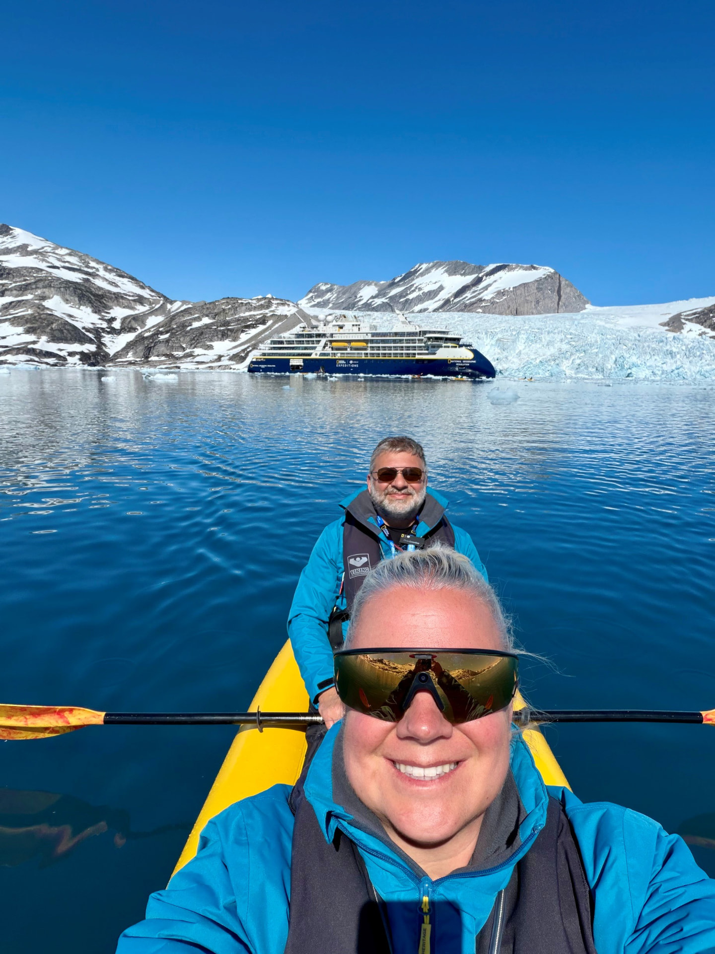 Two travelers in a yellow kayak paddling in front of a glacier and an expedition ship in Greenland